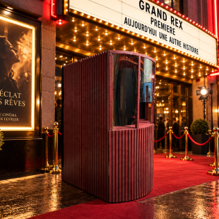 Caisse de cinéma vintage bordeaux mise en scène devant un cinéma de prestige avec tapis rouge